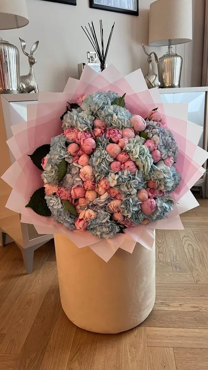 Pale blue hydrangeas and pink peonies wrapped in layered pink paper, standing on a velvet pouf