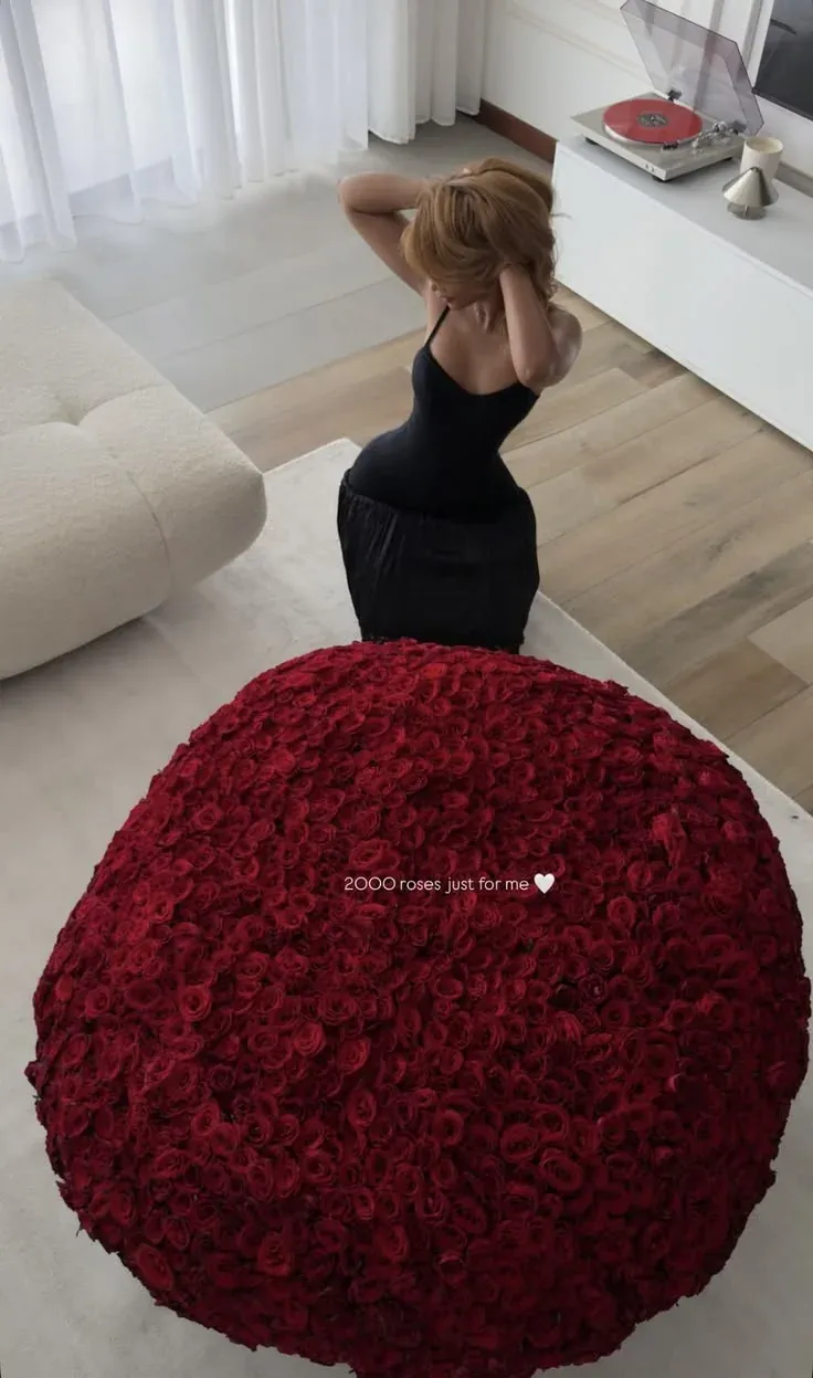 A huge dome of deep red roses on a living room rug, with a woman in a black dress kneeling behind it