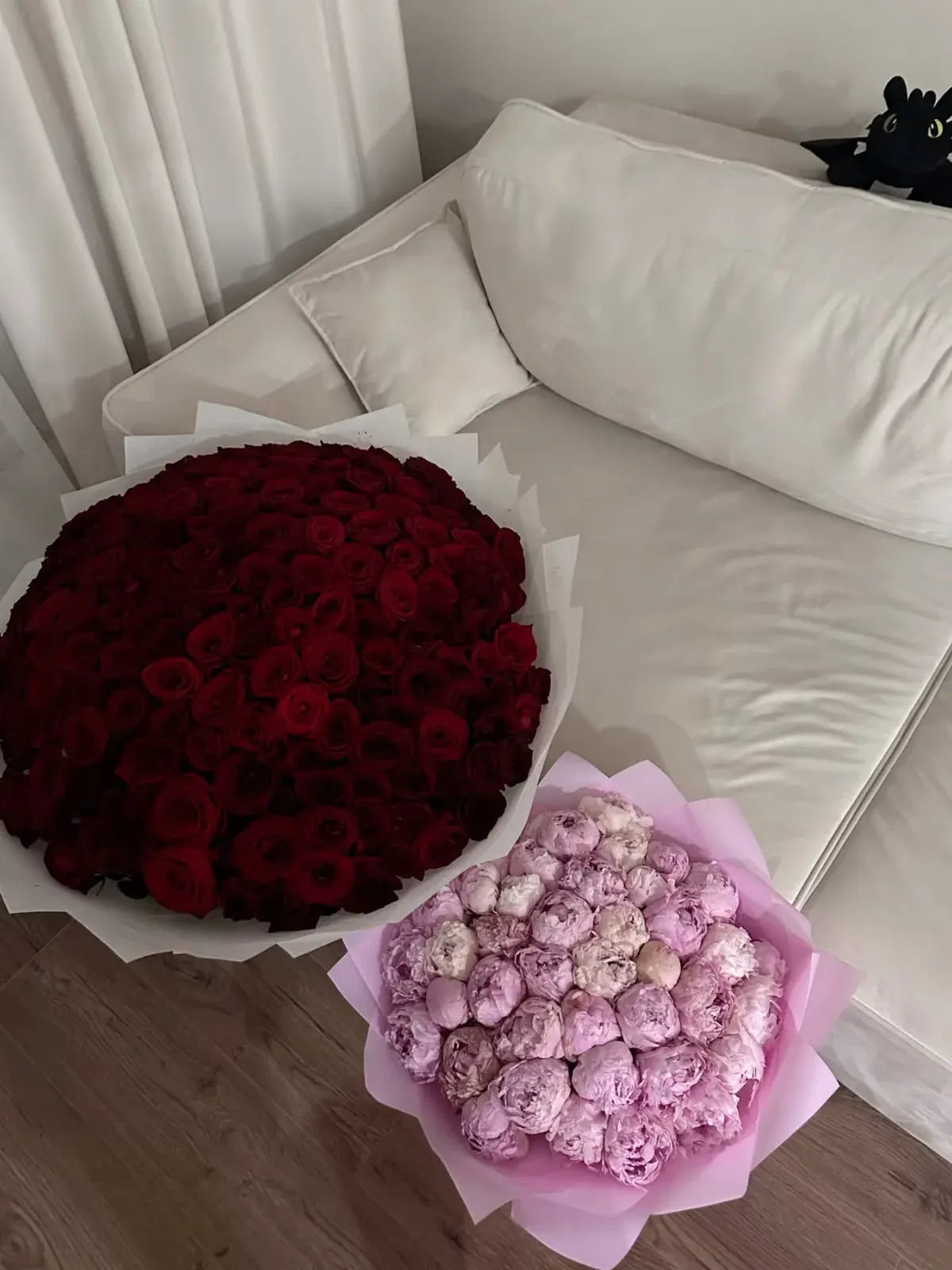 A large bouquet of red roses in white wrap beside a bouquet of pink peonies in pink wrap, set on a cream sofa