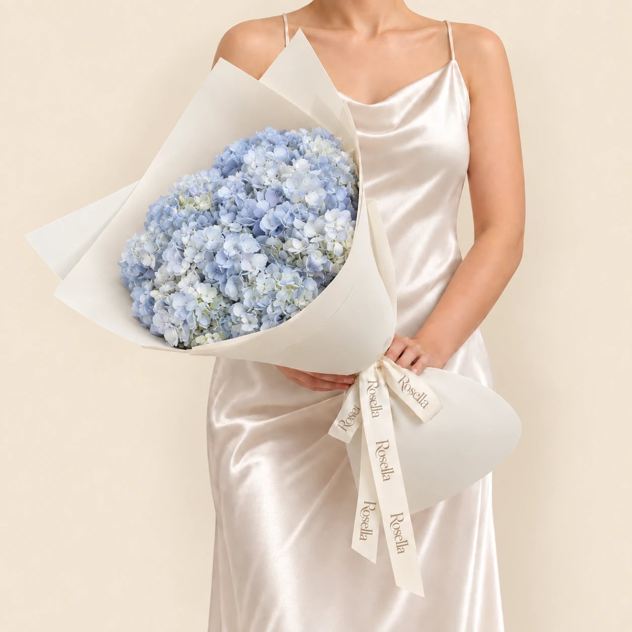 A bouquet of 12 pale blue hydrangeas wrapped in ivory paper and tied with a Rosella ribbon, held by a woman in a champagne satin dress