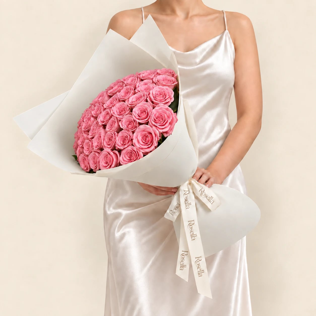 A bouquet of 50 pink roses wrapped in ivory paper and tied with a Rosella ribbon, held by a woman in a champagne satin dress