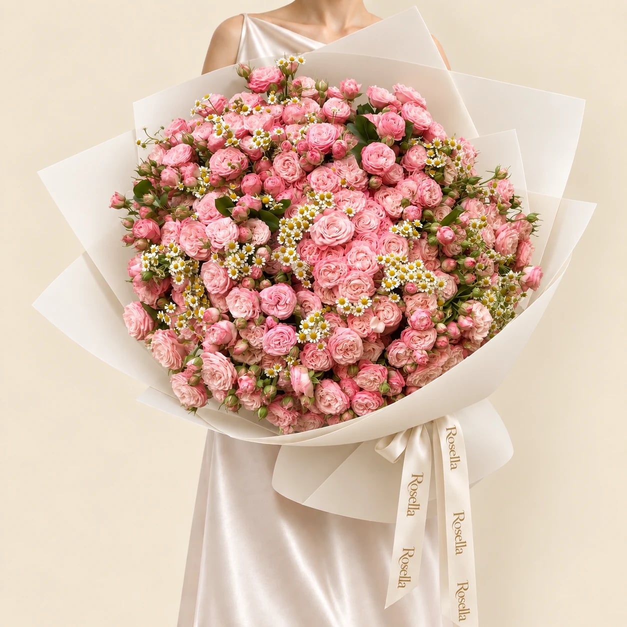 A huge dome of pink spray roses scattered with small white chamomile daisies in wide ivory wrap tied with a Rosella ribbon, held by a woman in a champagne satin dress