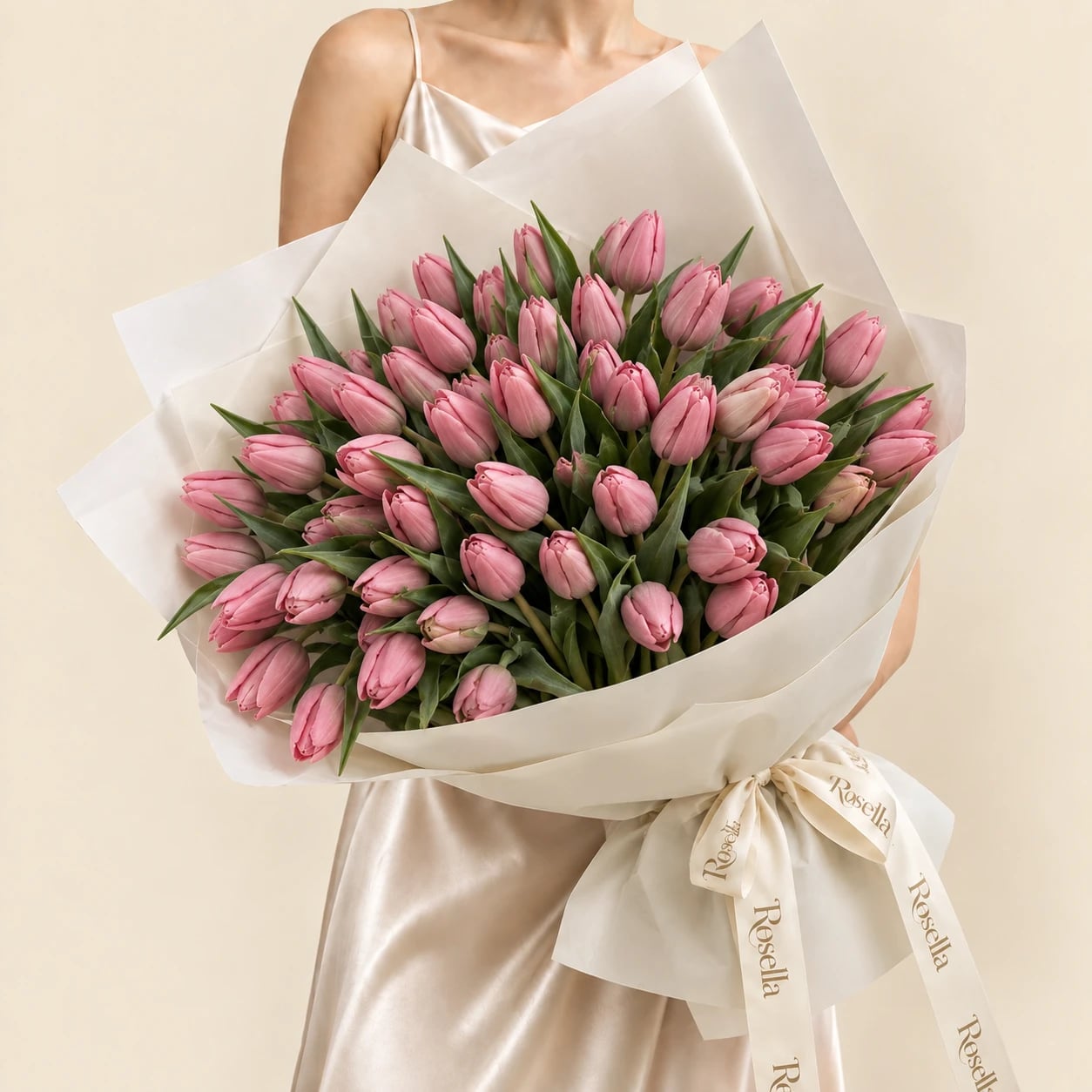 A bouquet of 50 soft pink tulips wrapped in ivory paper and tied with a large Rosella ribbon bow, held by a woman in a champagne satin dress