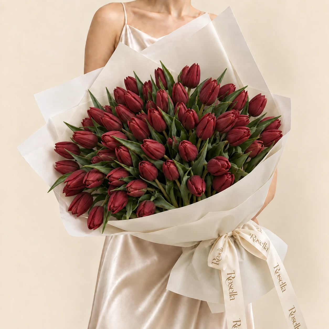 A bouquet of 50 deep red tulips wrapped in ivory paper and tied with a large Rosella ribbon bow, held by a woman in a champagne satin dress