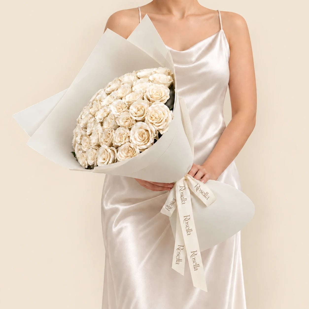 A bouquet of 50 white roses wrapped in ivory paper and tied with a Rosella ribbon, held by a woman in a champagne satin dress